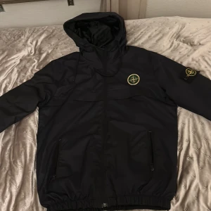 Svart jacka från Stone Island - Säljer en svart jacka från Stone Island med huva och dragkedja. Jackan har ett ikoniskt märke på armen och elastiska muddar vid ärmslut och nederkant. Perfekt för en stilren look.