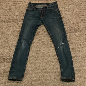 Blå jeans från Nudie Jeans - Säljer ett par snygga blå jeans från Nudie Jeans. De har en tight passform och slitna detaljer vid knäna för en cool look. Jeansen är tillverkade av 100% ekologisk bomull och har klassiska femficksdetaljer.