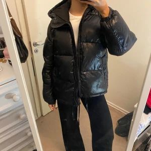 Svart pufferjacka med dragkedja - Säljer en svart pufferjacka med dragkedja. Jackan har en glansig yta och är perfekt för kyligare dagar. Den har långa ärmar och en hög krage för extra värme. Passar bra till både vardag och utflykter.