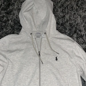 Ralph lauren zip hoodie - Säljer en stilren grå hoodie från Polo Ralph Lauren med dragkedja och klassisk logga på bröstet. Perfekt för en avslappnad look. Den har långa ärmar och en bekväm huva med snörning.