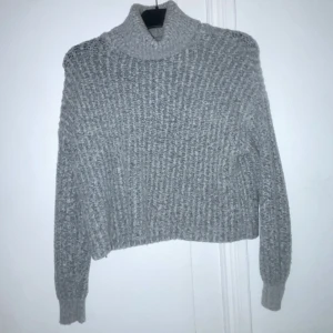 Grå stickad polotröja - Mysig grå stickad polotröja med en ribbad struktur. Perfekt för kyliga dagar med sin höga krage och långa ärmar. En klassisk och bekväm tröja för en stilren look.
