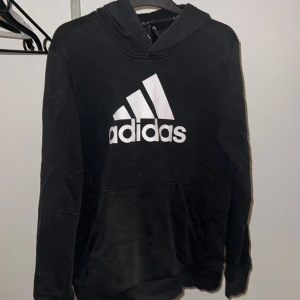 Svart hoodie från Adidas - Säljer en svart hoodie från Adidas med den klassiska loggan i vitt på framsidan. Tröjan har en stor ficka framtill och en bekväm huva. 