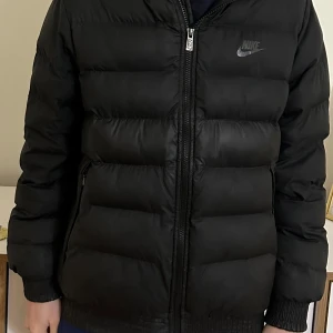 Svart dunjacka från Nike - Säljer en svart dunjacka från Nike med dragkedja och hög krage. Jackan har en pufferdesign och är perfekt för kyligare väder. Den har långa ärmar och två sidofickor med dragkedja. Jackan är i bra skick, inte använt så mycket. Pris kan alltid diskuteras . Storleken är 13-15 år alltså 158-170 cm!