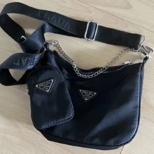 Svart axelväska  - Snygg svart axelväska med justerbar axelrem och kedjedetalj. Väskan har en dragkedja och en liten avtagbar väska framtill. Perfekt för att bära dina nödvändigheter med stil.