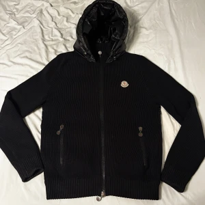 Moncler cardigan GAMLA MODELLEN!! - Snygg svart jacka från Moncler med ribbade ärmar och dragkedja framtill. Jackan har en quiltad huva och baksida, vilket ger en cool kontrast till det stickade materialet. Perfekt för kyligare dagar. Pris kan diskuteras för snabb affär. Inga stora problem. Logon är lite borta vid dragkedjan och vid fickorna men inget som man lägger märke till. 