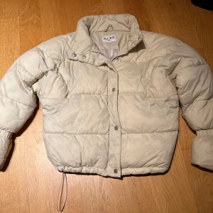 Beige pufferjacka från NA-KD - Säljer en beige pufferjacka från NA-KD i storlek 34. Jackan har en hög krage och stängs med dragkedja och knappar. Perfekt för kyliga dagar med sin quiltade design och elastiska ärmslut.