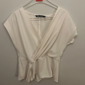Vit omlottopp från Zara - Säljer en stilren vit omlottopp från Zara. Toppen har en v-ringad design med knytning framtill och korta ärmar. Perfekt för en elegant och avslappnad look.