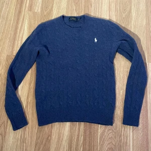 Blå merino/kashmir tröja från Polo Ralph Lauren - Säljer en stilren blå tröja från Polo Ralph Lauren i marino ull och kashmir. Tröjan har ett klassiskt kabelstickat mönster och en broderad logotyp på bröstet. Perfekt för kyliga dagar! Liten i storlek.