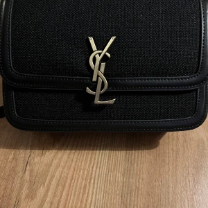 Svart axelväska från YSL  - Elegant svart axelväska från YSL med ikonisk logotyp i metall på framsidan. Väskan har en stilren design med detaljer i skinn och en praktisk storlek för vardagligt bruk. 