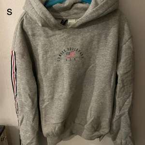 Grå hoodie med tryck - Säljer en grå hoodie med texten 'Sunset Boulevard USA' och en liten amerikansk flagga på bröstet. Tröjan har långa ärmar med randiga detaljer i rött, vitt och blått längs ärmarna. Perfekt för en avslappnad stil.
