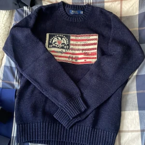 Mörkblå stickad tröja från Polo Ralph Lauren - Säljer en snygg mörkblå stickad tröja från Polo Ralph Lauren med ett coolt amerikanskt flaggmotiv och årtalet 1967 på bröstet. Tröjan har långa ärmar och ribbade muddar vid ärmslut och nederkant. Perfekt för en stilren look!