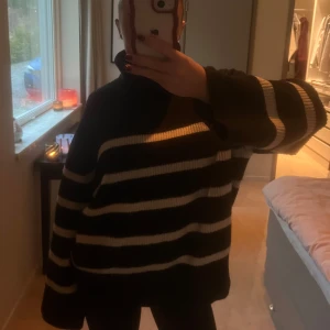 Svart och vit randig stickad tröja - Mysig svart stickad tröja med breda vita ränder. Tröjan har en hög krage och långa ärmar, perfekt för kyliga dagar. Den har en avslappnad passform som ger en bekväm känsla.