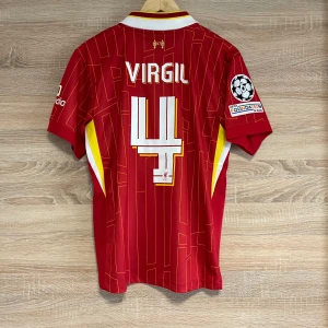 Liverpool Hemma Virgil 4 XL - Snygg röd fotbollströja från Nike med Virgil och nummer 4 på ryggen. Tröjan har korta ärmar och ett coolt mönster i gult. Den har även UEFA Foundation-märke på ärmen och L.F.C.-logga på bröstet. Perfekt för fotbollsfans!