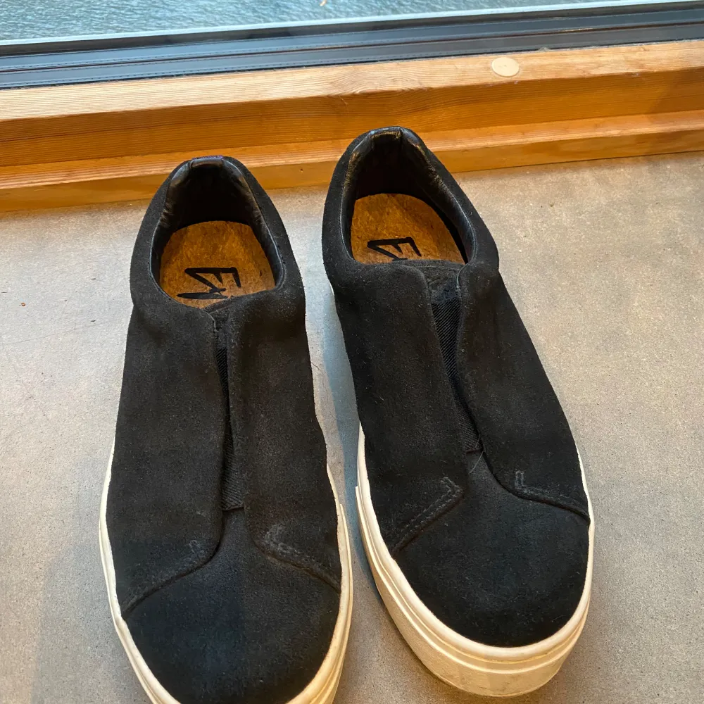 Snygga svarta sneakers i mocka med vit sula. De har en stilren design utan snörning, vilket gör dem enkla att ta på och av. Perfekta för en avslappnad stil.. Kengät.