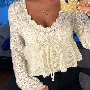 Vit stickad tröja med knytband - Charmig vit stickad tröja med volangdetaljer och knytband i midjan. Tröjan har en v-ringad hals och långa armar. 