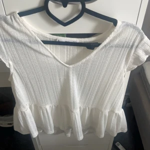 Vit blus - Säljer en söt vit ribbad blus med korta ärmar och volangdetalj i nederkant. Blusen har en v-ringad hals och är perfekt för en somrig look.