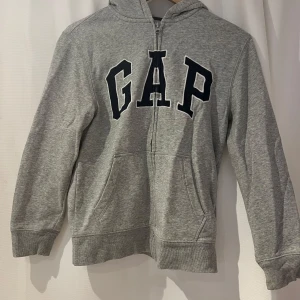 Grå hoodie från GAP Kids - Säljer en klassisk grå hoodie från GAP Kids med dragkedja och stor logga på framsidan. Stl Kids XL/150-152. Några svaga blå fläckar: en uppe på huvan (framtill), en på ena mudden för handleden (baksida), se bilder. Annars bra skick. 