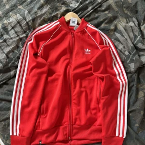 Röd zip tröja Adidas - Säljer en snygg röd zip tröja från Adidas med de klassiska tre vita ränderna längs ärmarna. Tröjan har en dragkedja framtill och är långärmad. Perfekt för träning eller som en stilren vardagsjacka.