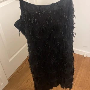Svart klänning med paljetter och fransar - Säljer en superfin svart klänning med massor av paljetter och fransar som ger en riktigt festlig look. Smala axelband och en tight passform som framhäver figuren. Aldrig använd. Storlek M men liten i storleken 