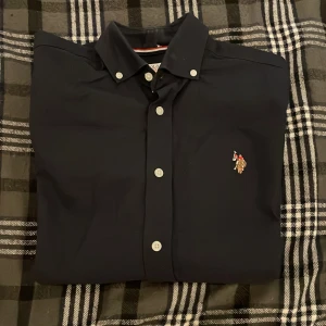US Polo Assn Skjorta - Fin US Polo Assn Skjorta i marinblå färg. Den är i bra skick 8,7/10 och är fin till fester/kalas/partyn/dejter mm. Säljer den för att den inte passar. Har inte använt så mycket för skaffade en ny skjorta. Pris kan diskutera. 