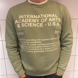 Grön sweatshirt med tryck - Säljer en grön sweatshirt med tryck från International Academy of Arts & Science - U.S.A. Tröjan har text i vitt framtill och är perfekt för en avslappnad stil. Den är långärmad och gjord i ett mjukt material som är skönt att ha på sig. Perfekt för höst och vår!