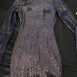 Glittrig klänning med öppen rygg - Säljer en superfin glittrig klänning i mörkblått med långärmad design. Den har en snygg öppen rygg som verkligen sticker ut. Perfekt för fest eller nyår! Klänningen är tight och har ett glittrigt mönster. Ända problemet är att lite av det glittriga mönster har gått av som ni ser 