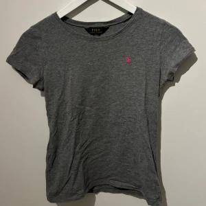 Grå t-shirt från Polo Ralph Lauren - Säljer en stilren grå t-shirt från Polo Ralph Lauren med en liten rosa logga på bröstet. T-shirten är kortärmad och tillverkad i mjukt bomullsmaterial. Perfekt för en avslappnad look.