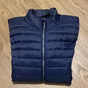 Mörkblå pufferjacka - Säljer en stilren mörkblå pufferjacka med dragkedja framtill. Jackan har långa ärmar och två praktiska fickor med dragkedjor. Perfekt för kyligare dagar.