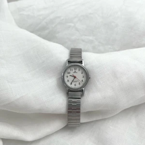 Silverfärgad klocka med vit urtavla - Klockan fungerar.  Klockan Mireille från märket Lorus är ett elegant armbandsur med en vit urtavla och tydliga svarta siffror. Klockan har ett silverfärgat metallarmband som ger en tidlös look. Perfekt för den som söker en stilren och funktionell klocka. Längd/yttre omkrets på klockan:20cm  Urtavla: 20mm/2cm