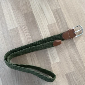 Grönt flätat bälte med bruna detaljer - Snyggt grönt flätat bälte med bruna läderdetaljer och silverfärgat spänne. Perfekt för att ge en stilren touch till din outfit. Bältet har en klassisk design med en modern twist.