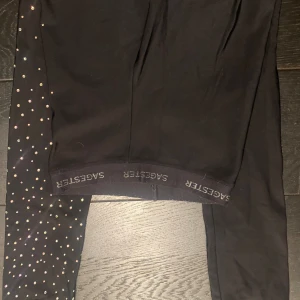 Svarta leggings med glittriga detaljer från SAGESTER - Snygga svarta leggings från SAGESTER med glittriga detaljer på ena benet. Perfekta för att ge en extra touch till din outfit. De har en bekväm passform och är idealiska för både vardag och speciella tillfällen.