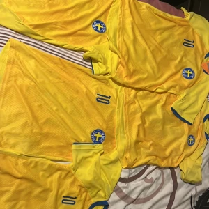 Gul fotbollströja med nummer 10 - Säljer en gul fotbollströja med blå detaljer och nummer 10. Tröjan har korta ärmar och Sveriges emblem på bröstet. Perfekt för fotbollsfans!