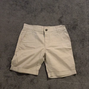 Beige shorts med knappdetaljer - Snygga beige shorts med knappstängning fram och två bakfickor med knappar. Perfekta för en avslappnad stil. De har en klassisk passform och är gjorda i ett bekvämt material.