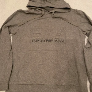 Grå hoodie från Emporio Armani EA7 - Säljer en stilren grå hoodie från Emporio Armani med  EA7 logga på bröstet. Tröjan har en klassisk design med huva och känguruficka framtill. 