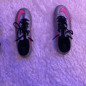 Nike fotbollsskor i silver och rosa - Snygga fotbollsskor från Nike i en cool silverfärg med rosa detaljer och svarta snören. Skorna har en modern design med dobbar för bra grepp på planen. Perfekta för fotbollsspelare som vill sticka ut.
