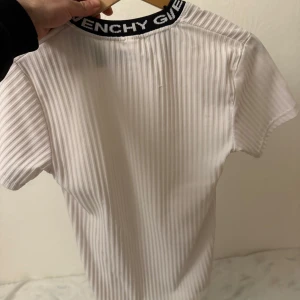 Vit ribbad t-shirt från Givenchy - Säljer en stilren vit ribbad t-shirt från Givenchy med korta ärmar. Den har en svart krage med Givenchy-loggan i vitt. Perfekt för en trendig och avslappnad look.