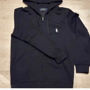 Svart hoodie från Ralph Lauren - Säljer en stilren svart hoodie från Ralph Lauren med dragkedja och det klassiska logotypbroderiet på bröstet. Perfekt för en avslappnad look. Den har en bekväm passform och är tillverkad i mjukt material.