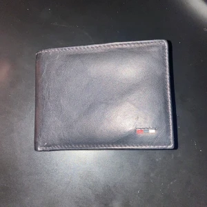 Svart plånbok från Tommy Hilfiger - Snygg svart plånbok i skinn från Tommy Hilfiger. Den har flera fack för kort och en stilren design med en liten logotyp i hörnet. Perfekt för att hålla ordning på dina kort och kontanter.