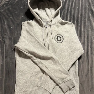 Grå hoodie från DirtCült - Säljer en grå hoodie från DirtCült med dragkedja och tryck på både fram- och baksidan. Den har en justerbar huva med snören och en praktisk ficka framtill. Perfekt för en avslappnad stil. 