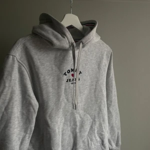 Grå hoodie från Tommy Jeans - Säljer en stilren grå hoodie från Tommy Jeans med logotyp på bröstet. Hoodien har en justerbar dragsko i huvan och ribbade muddar vid ärmslut och nederkant. Perfekt för en avslappnad look. Använd fåtal gånger!