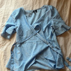 Blå omlottopp med prickigt mönster - Säljer en söt blå omlottopp från Vero Moda med vita prickar. Toppen har trekvartsärmar och knytband i midjan för en justerbar passform. Perfekt för vår och sommar!