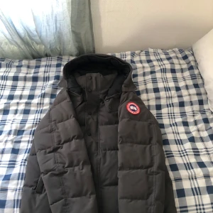 Grå Canada Goose jacka priset kan sänkas under snabb affär  - Säljer en svart dunjacka från Canada Goose med huva och ett klassiskt märke på ärmen. Jackan är perfekt för kalla dagar och har en stilren design med dragkedja och knappar framtill använd i 2/3 månader.