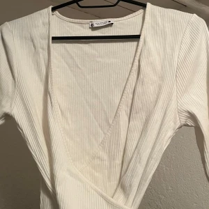 Vit ribbad omlottopp från Urban Outfitters - Säljer en vit ribbad omlottopp från zara. Toppen har en djup v-ringning och kan knytas med band runt om. Nästan aldrig använd storlek M men passar nog s också💕
