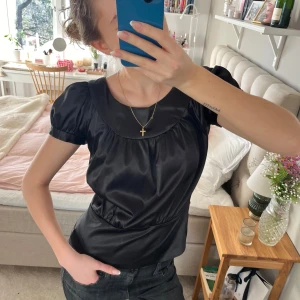 Svart satinblus - Snygg svart blus i satin med korta ärmar och rund hals. Blusen har en elegant glans och knäppdetaljer på ärmarna som ger en stilren look. Perfekt för att lyfta vilken outfit som helst 💕💕