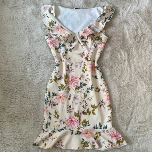 Blommig klänning med volangdetaljer - Charmig blommig klänning i ljusa toner med volangdetaljer vid axlar och fåll. Klänningen har en v-ringad design och är ärmlös, vilket ger en somrig känsla. Perfekt för varma dagar och speciella tillfällen.