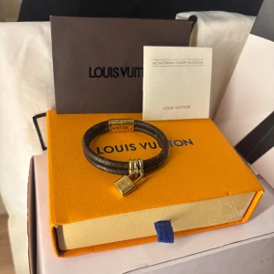 Brunt armband med lås från Louis Vuitton - Elegant brunt armband från Louis Vuitton med ett guldfärgat lås och detaljer. Armbandet har en stilren design med märkets ikoniska monogram. Perfekt accessoar för att lyfta din stil. Storlek 17cm