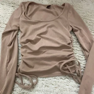 Beige ribbad långärmad topp - Säljer en beige ribbad topp med långa ärmar och knytning i sidorna. Perfekt för en stilren och bekväm look. Passar bra till både jeans och kjol.