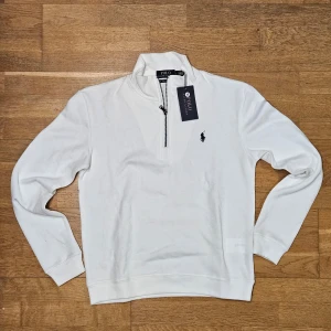Vit tröja från Polo Ralph Lauren Quarter zip - Säljer en stilren vit tröja från Polo Ralph Lauren med dragkedja vid halsen och det klassiska logotypbroderiet på bröstet. Tröjan har långa ärmar och en bekväm passform. Perfekt för en avslappnad och snygg look.