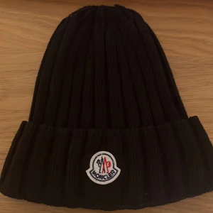 Svart ribbad mössa från Moncler - PRIS diskuteras vid snabb affär!!Snygg svart ribbad mössa från Moncler med broderad logotyp framtill. Perfekt för kyliga dagar och ger en stilren look. Tillverkad i ett mjukt och bekvämt material.