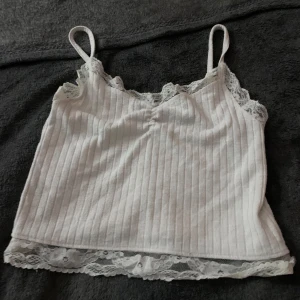 Vit ribbad linnetopp från FB Sister - Säljer en söt vit ribbad linnetopp från FB Sister med spetsdetaljer vid halsringningen och nederkanten. Perfekt för varma dagar eller som ett lager under en tröja. Den har smala axelband och en bekväm passform(den har ett håll i ryggen som är lite synligt men lätt att laga)